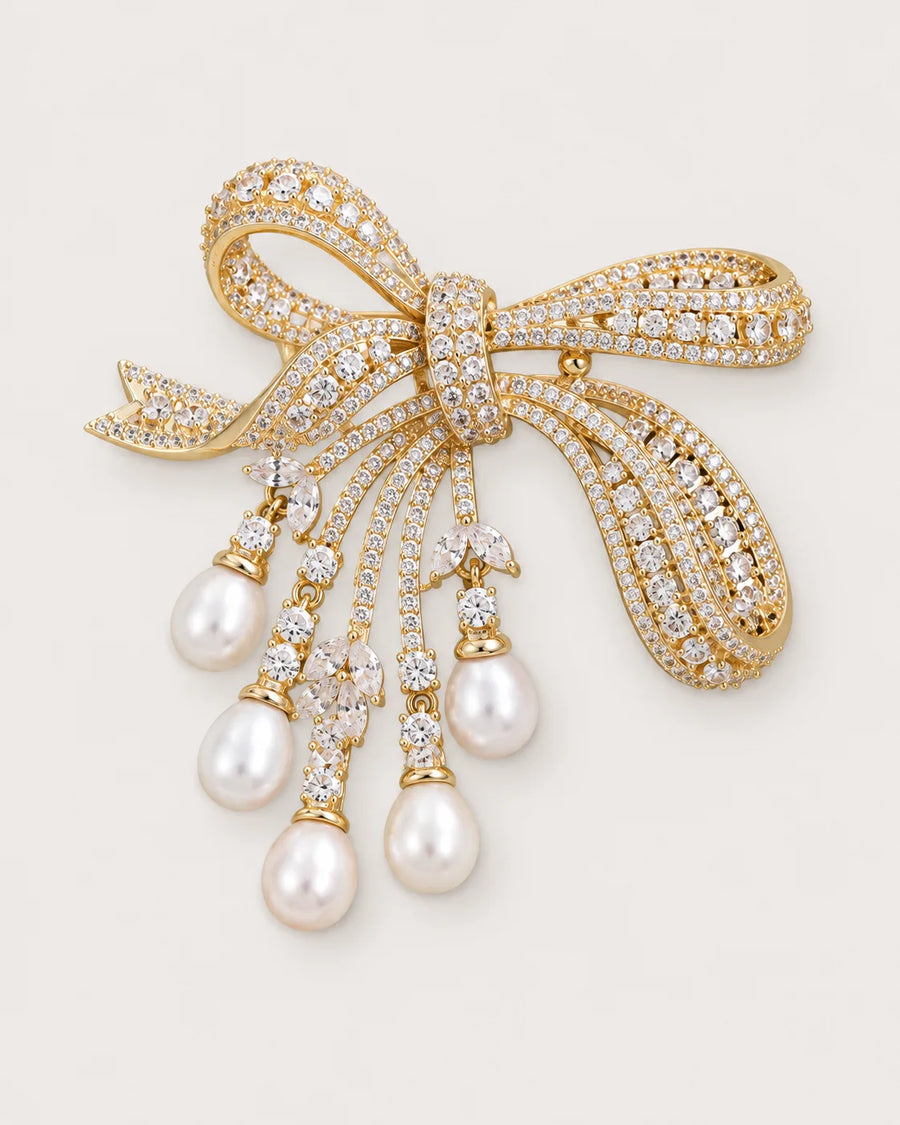 Gold-tone bow brooch with five freshwater pearl drops on a warm neutral background