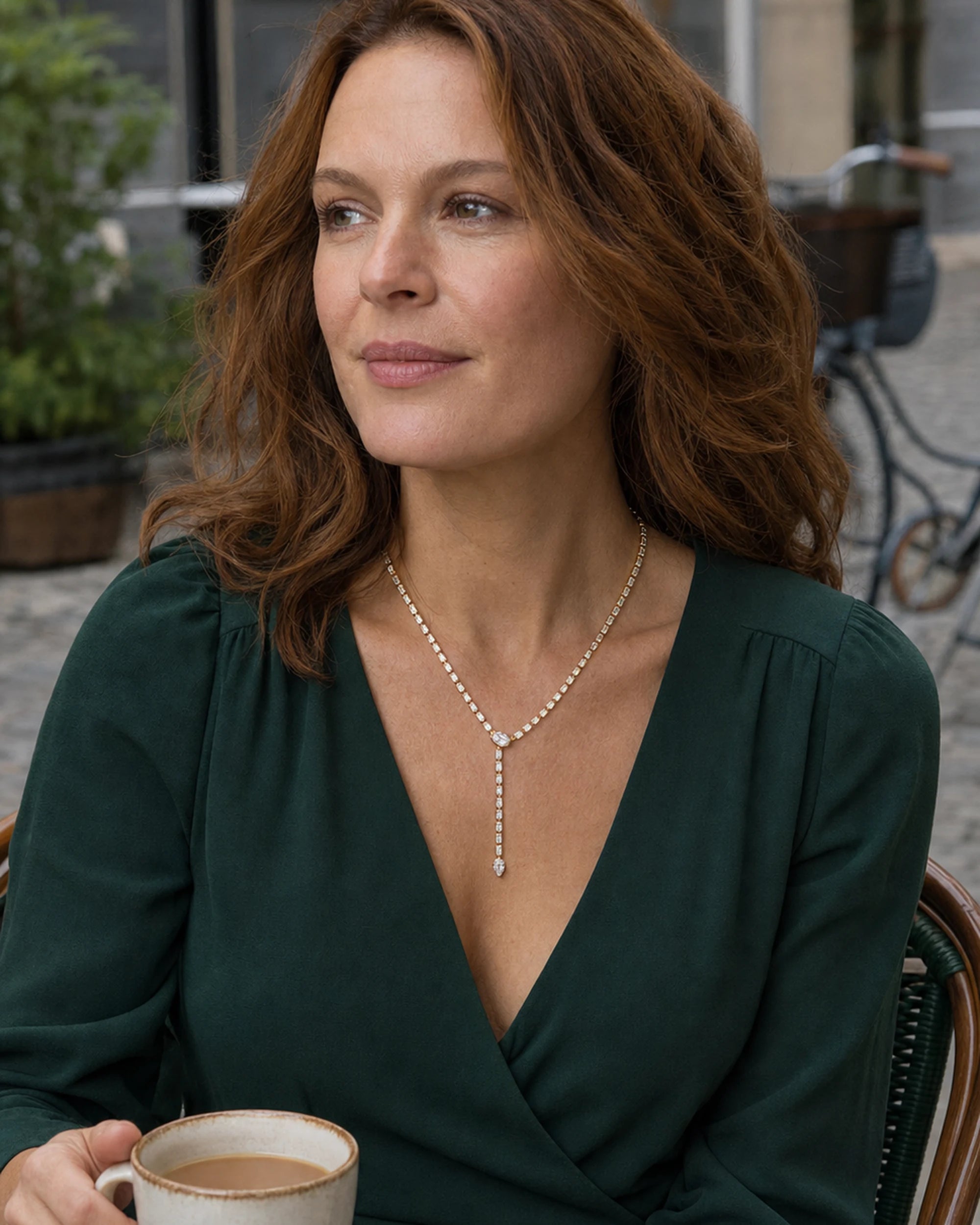 Red-haired woman wearing the Liora Gold Crystal Y-Drop Necklace with a green wrap dress at a café
