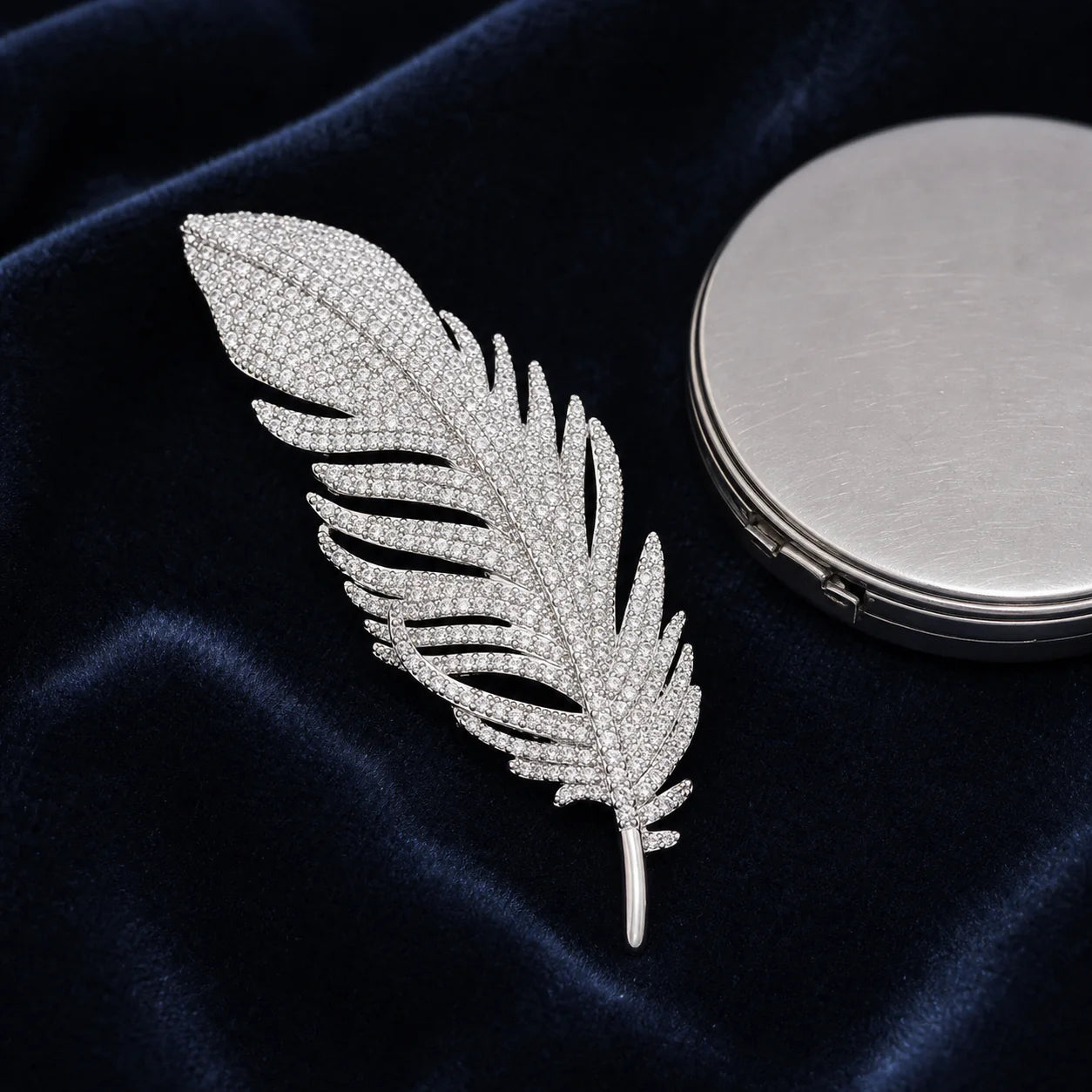 Lumi Crystal Feather Brooch displayed on midnight-blue velvet beside a silver compact