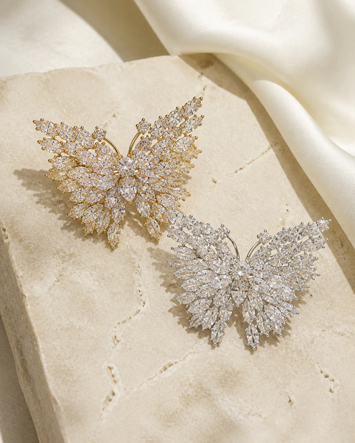 Gold-tone and silver-tone Eveli Butterfly Brooch variants arranged on pale limestone and ivory silk