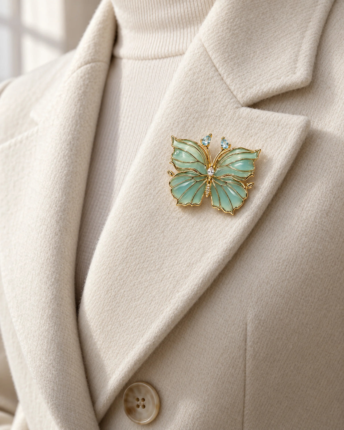 Ilma blue-green enamel butterfly brooch pinned within an ivory wool coat lapel.
