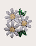 Ilona silver-tone daisy trio brooch with gold-tone centers and green leaves on a warm neutral background
