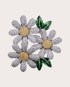 Ilona silver-tone daisy trio brooch with gold-tone centers and green leaves on a warm neutral background
