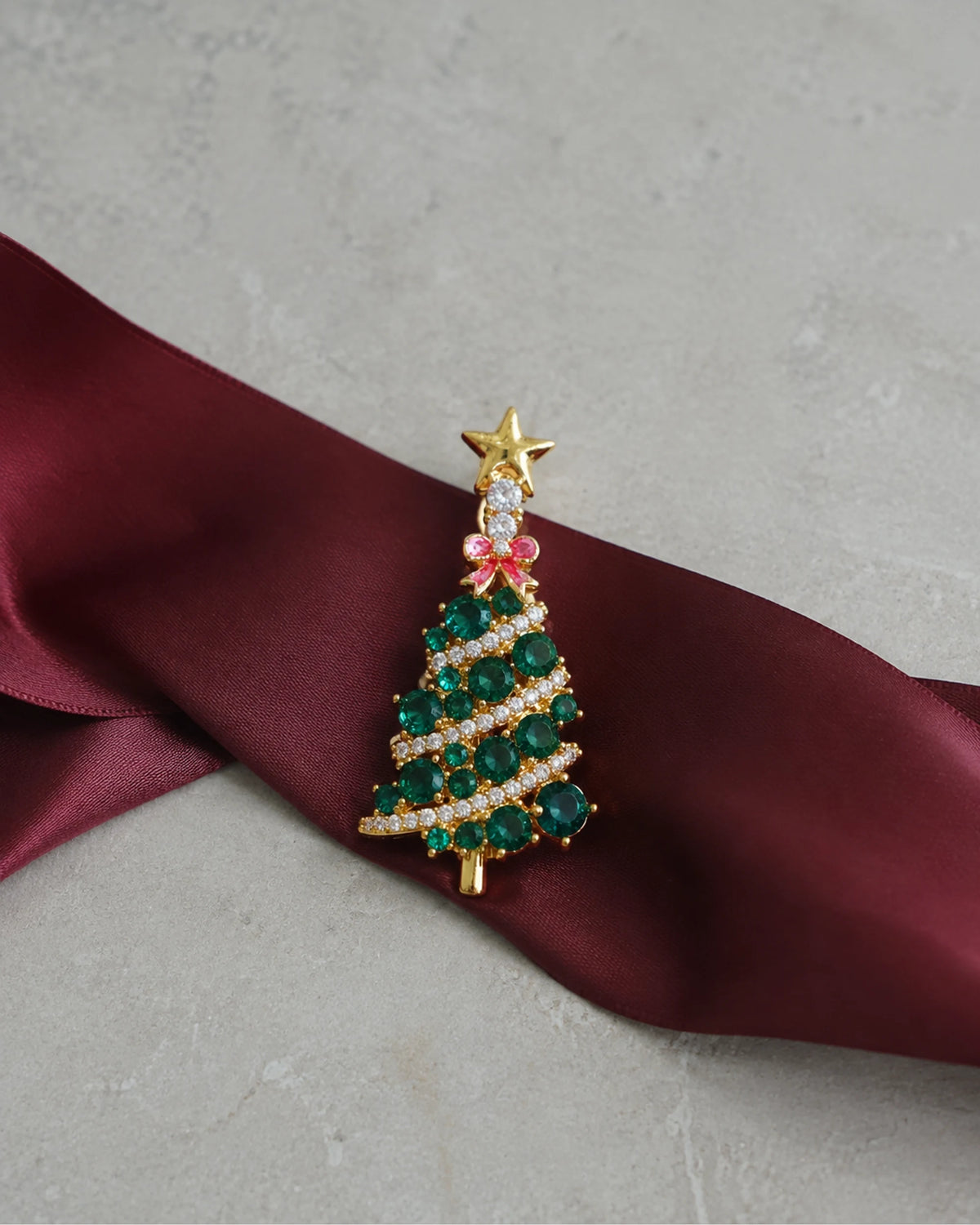 Green tree brooch resting on burgundy ribbon over pale stone.