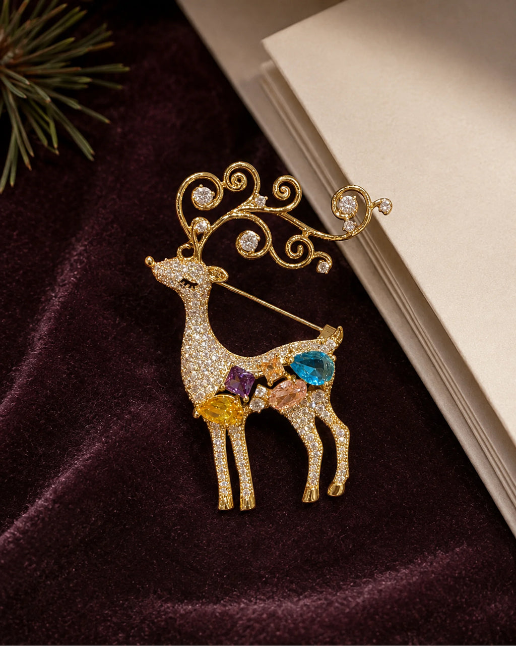 Niva deer brooch on plum fabric beside a blank ivory card and evergreen detail.