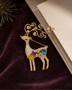 Niva deer brooch on plum fabric beside a blank ivory card and evergreen detail.