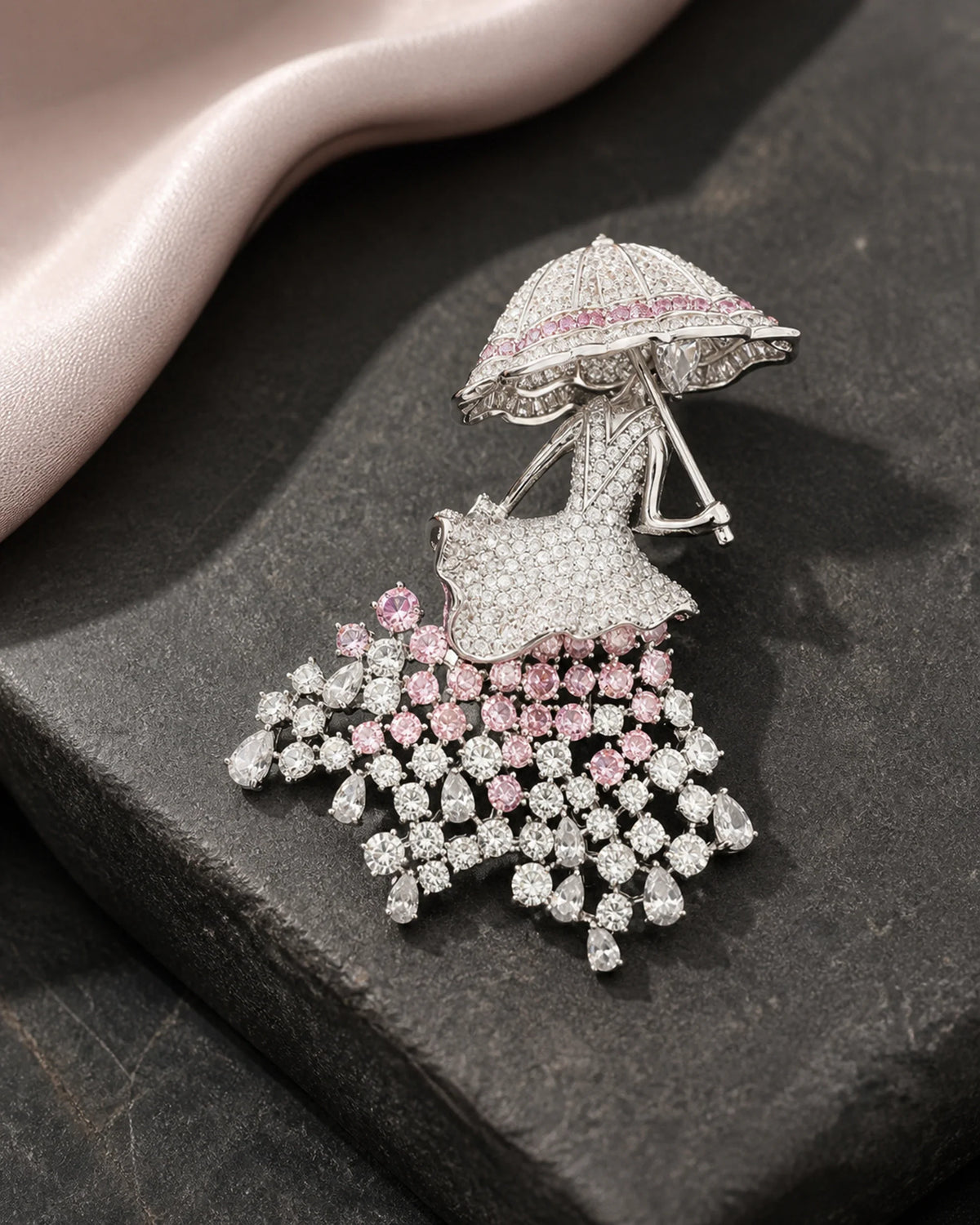 Noora parasol lady brooch resting on charcoal stone beside blush silk