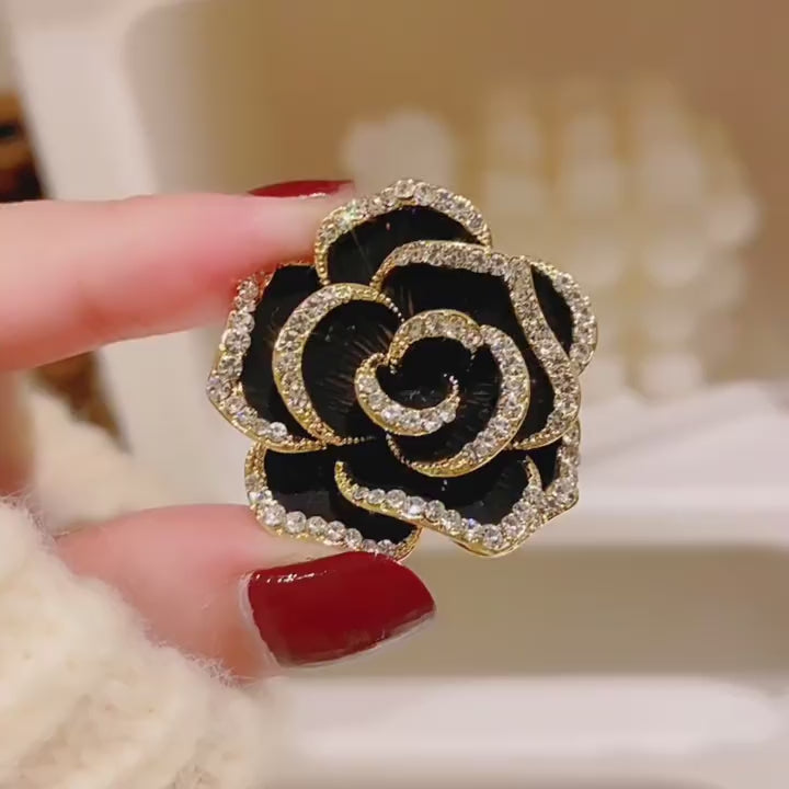 Noir black camellia brooch held in a real hand, showing glossy ribbed petals and crystal borders in motion