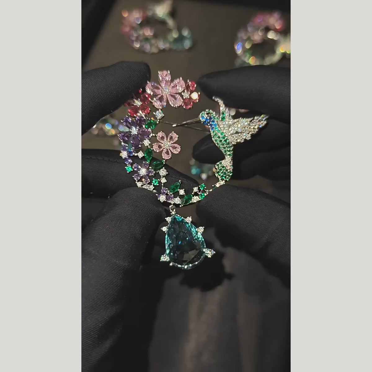 Pihla hummingbird floral wreath brooch held in black gloves, showing colorful flowers, green leaves and a blue pear-shaped drop in motion.