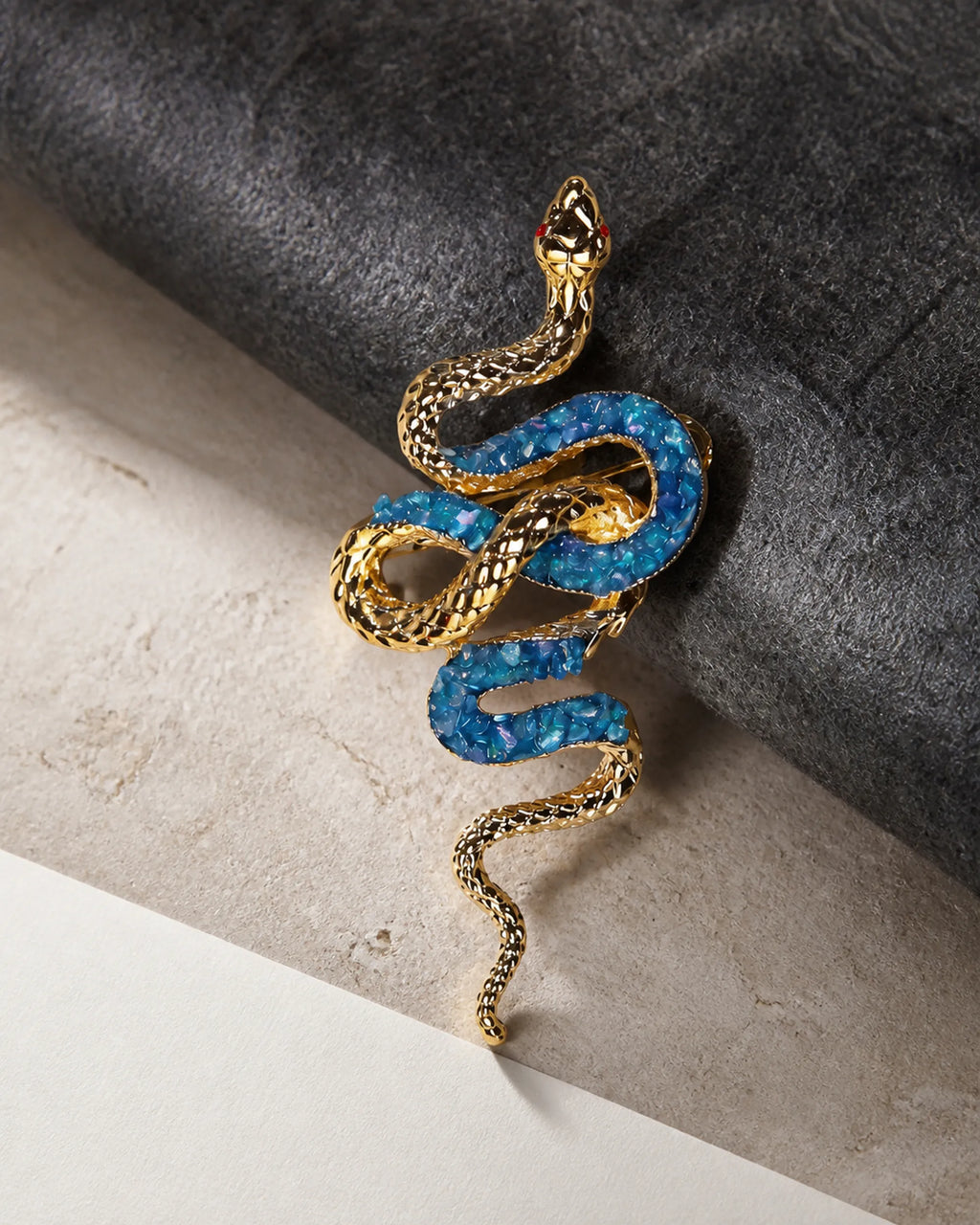 Sisu gold and lake-blue snake brooch resting on charcoal wool, pale limestone and ivory paper