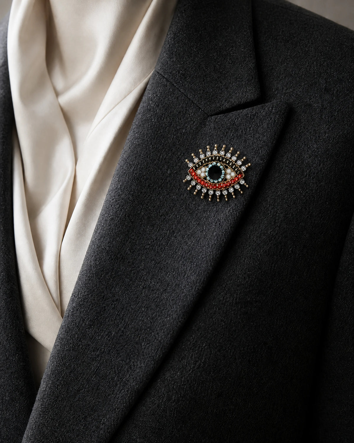 Close view of the Suoja eye brooch pinned fully to a folded charcoal blazer lapel at its true approximately 4.0 by 3.2 centimetre scale