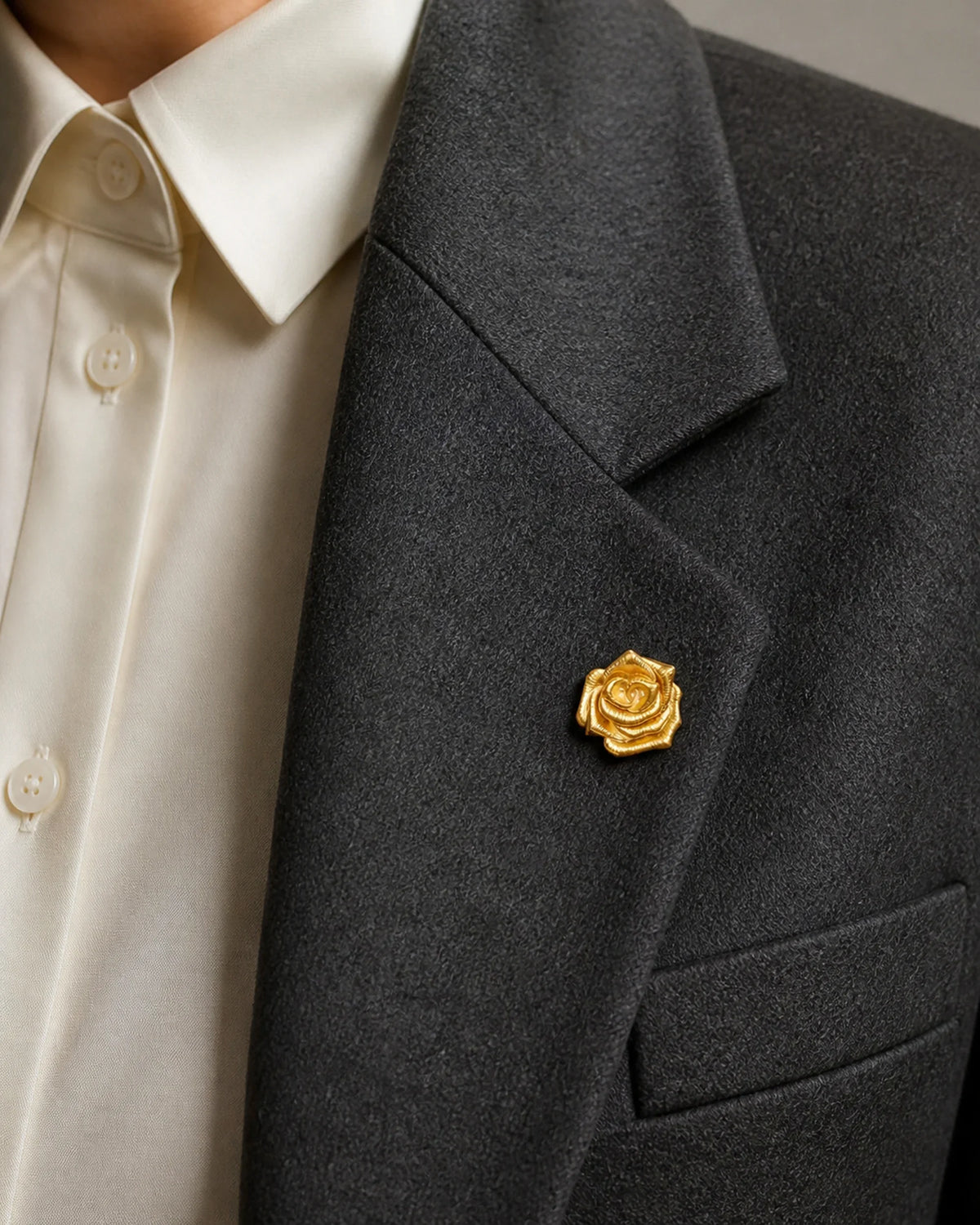 Close view of the 2.2 cm Taimi Mini Gold Camellia Brooch pinned to the folded lapel of a charcoal blazer