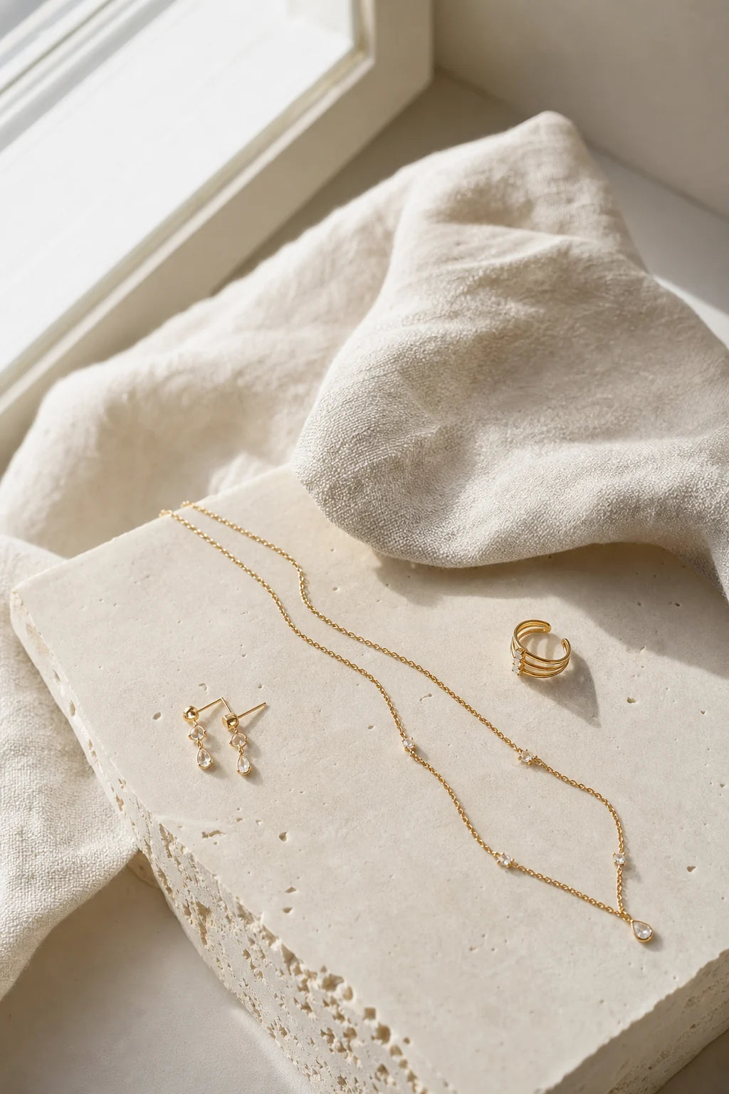 Delicate gold jewelry arranged on pale stone and linen
