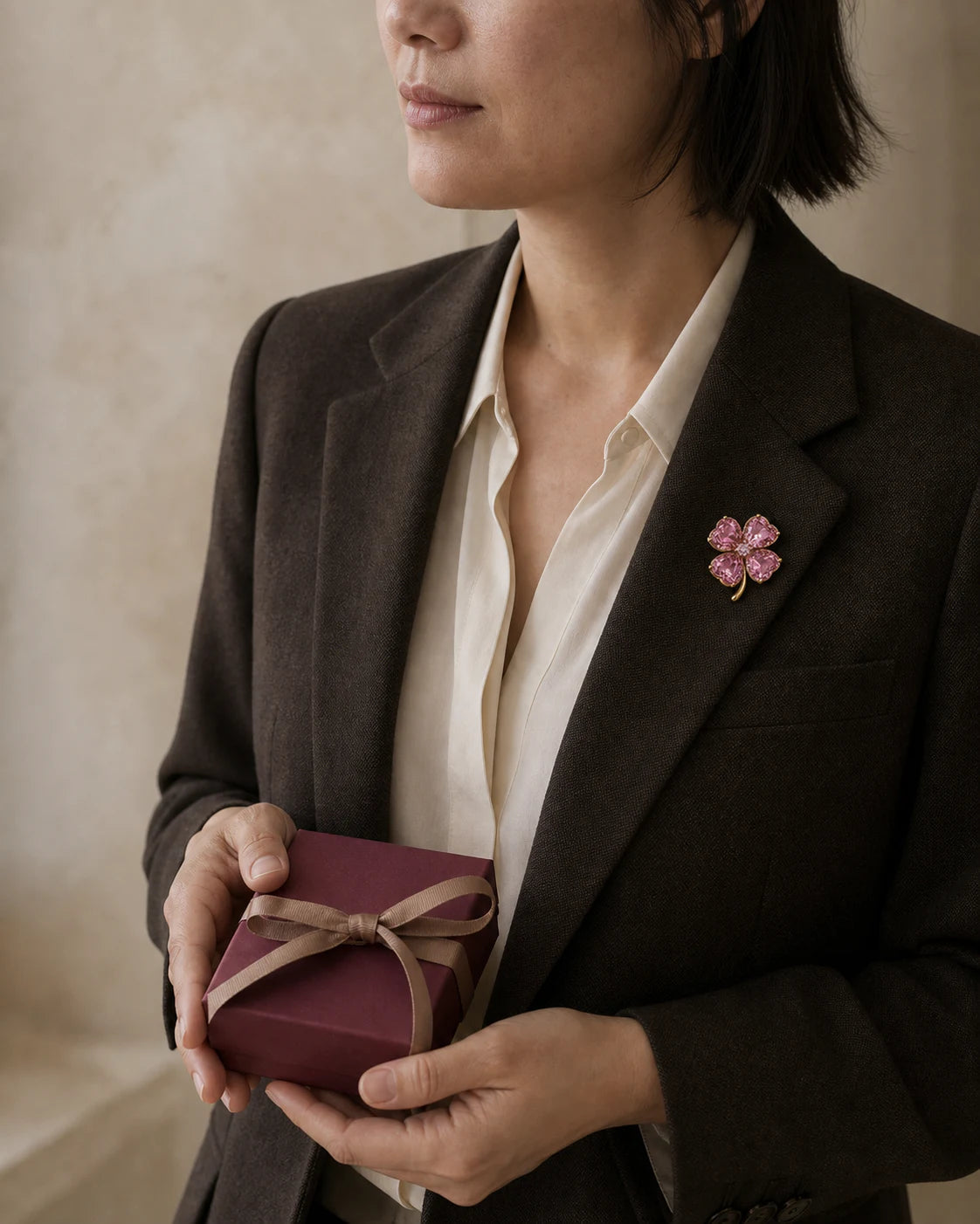 Woman in an espresso blazer wearing the pink Lykke Clover Brooch and holding a burgundy gift box